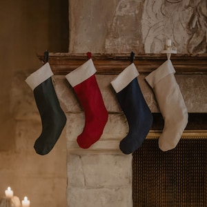May include: Four Christmas stockings hanging on a fireplace mantel. The stockings are made of fabric and are in the colors red, green, blue, and beige.