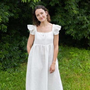 May include: A floor-length white dress featuring a square neckline, flutter sleeves, and a gathered waistline. The garment is constructed with a simple bodice and a loose, flowing skirt that extends to the ankles, providing a clean and neutral appearance.