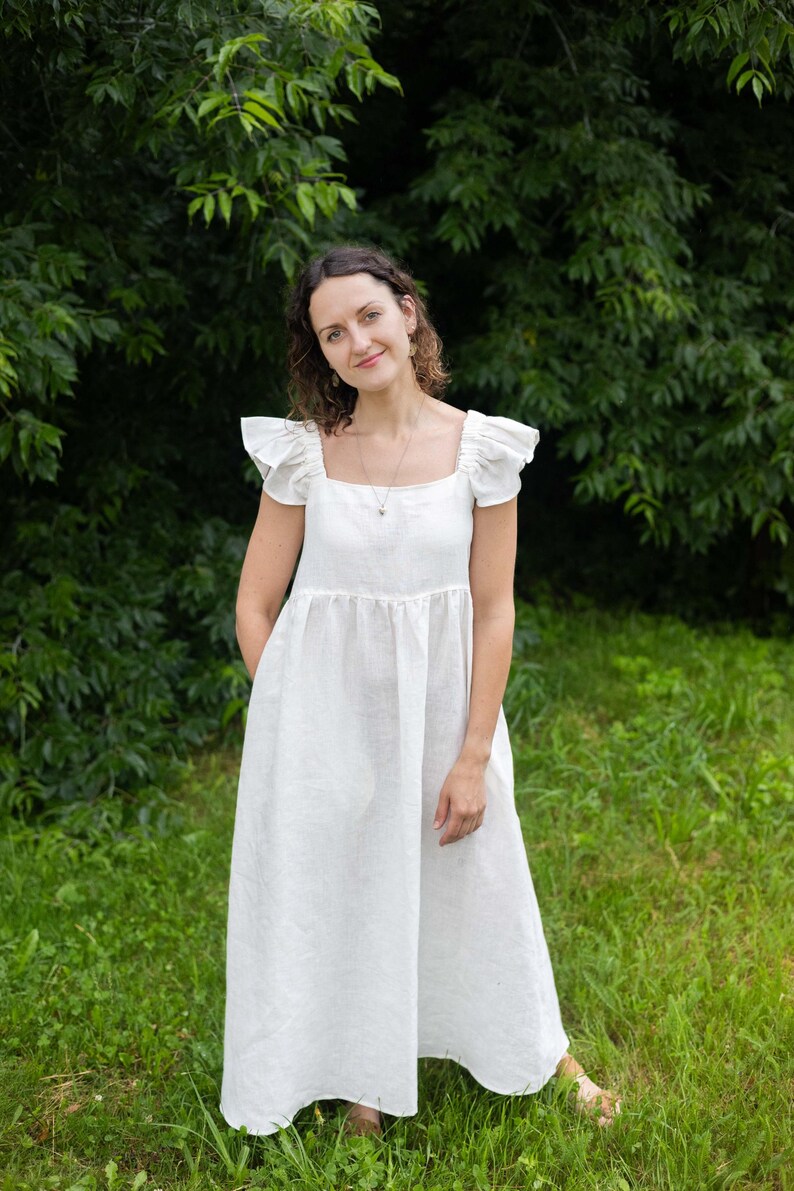 May include: A floor-length white dress featuring a square neckline, flutter sleeves, and a gathered waistline. The garment is constructed with a simple bodice and a loose, flowing skirt that extends to the ankles, providing a clean and neutral appearance.