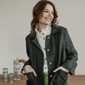 Pode incluir: Uma mulher vestindo uma jaqueta de linho verde escura com uma camisa branca de babados e calças verdes. Ela está em pé em frente a uma mesa de madeira com uma garrafa de vidro e copos em cima.