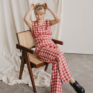 May include: A woman wearing a red and white gingham jumpsuit sits on a wooden chair with a woven cane seat. She is wearing black shoes and has her blonde hair pulled back.