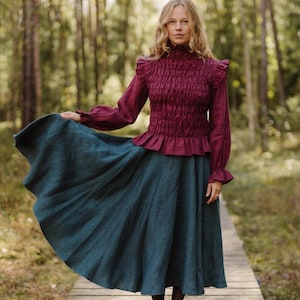 May include: A long-sleeved burgundy blouse with a high smocked neckline, ruffled shoulders, and flared cuffs, paired with a dark teal midi skirt. The blouse features a textured, gathered bodice design, while the skirt has a full, flowing silhouette.