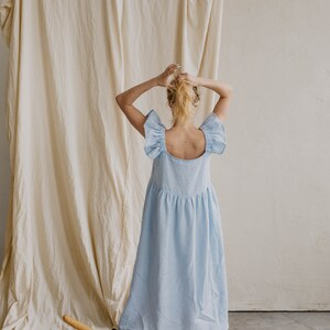 May include: A light blue midi dress featuring a square neckline, wide ruffled flutter sleeves, and a gathered waistline. The dress has a loose, flowing silhouette that falls below the knees, crafted from a solid-colored fabric with a visible texture.