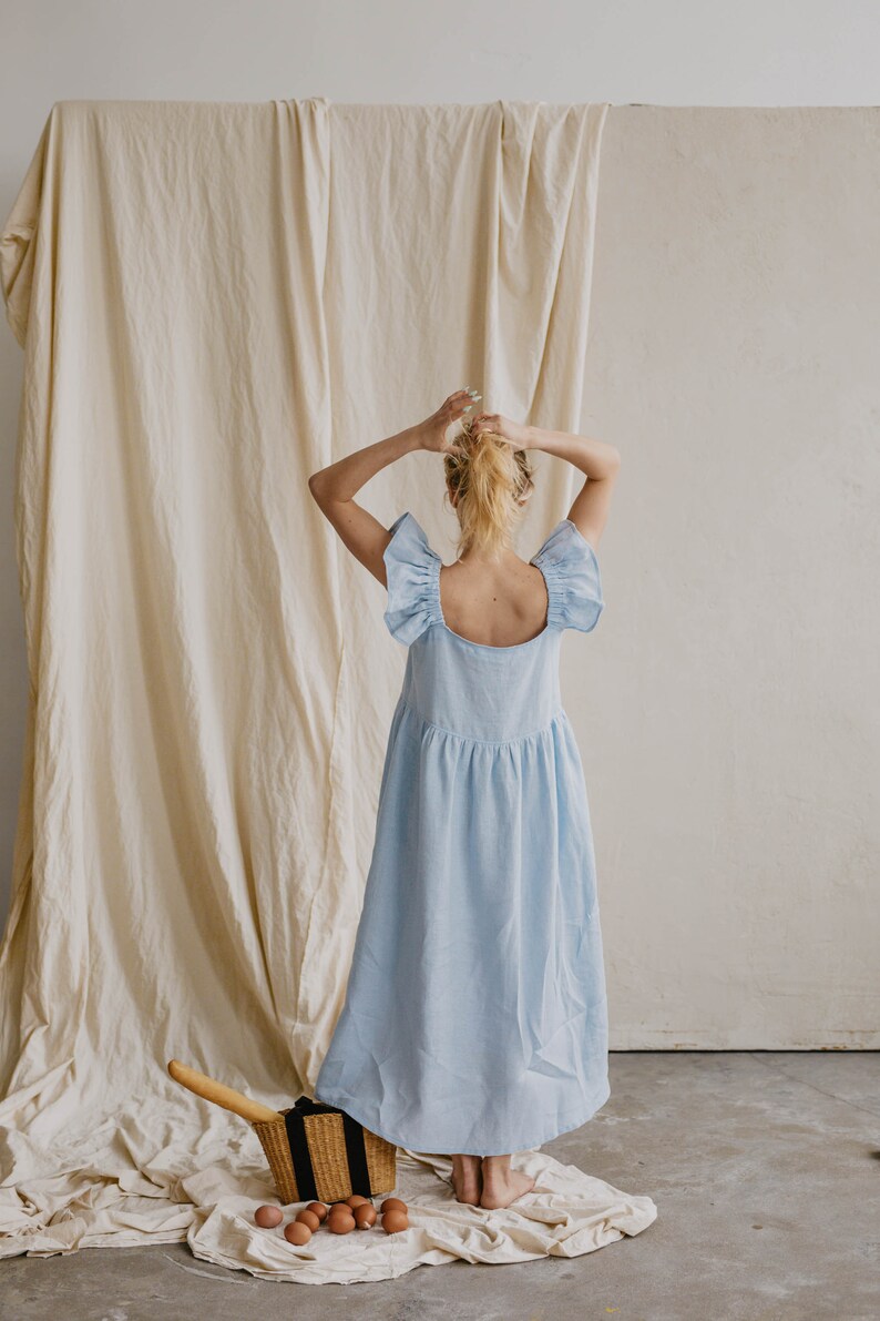 May include: A light blue midi dress featuring a square neckline, wide ruffled flutter sleeves, and a gathered waistline. The dress has a loose, flowing silhouette that falls below the knees, crafted from a solid-colored fabric with a visible texture.