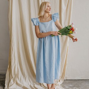 May include: A light blue midi dress featuring a square neckline and ruffled cap sleeves. The dress has a relaxed, loose-fitting silhouette that falls below the knees. The garment is made of a solid-colored fabric and is held with a bouquet of tulips.