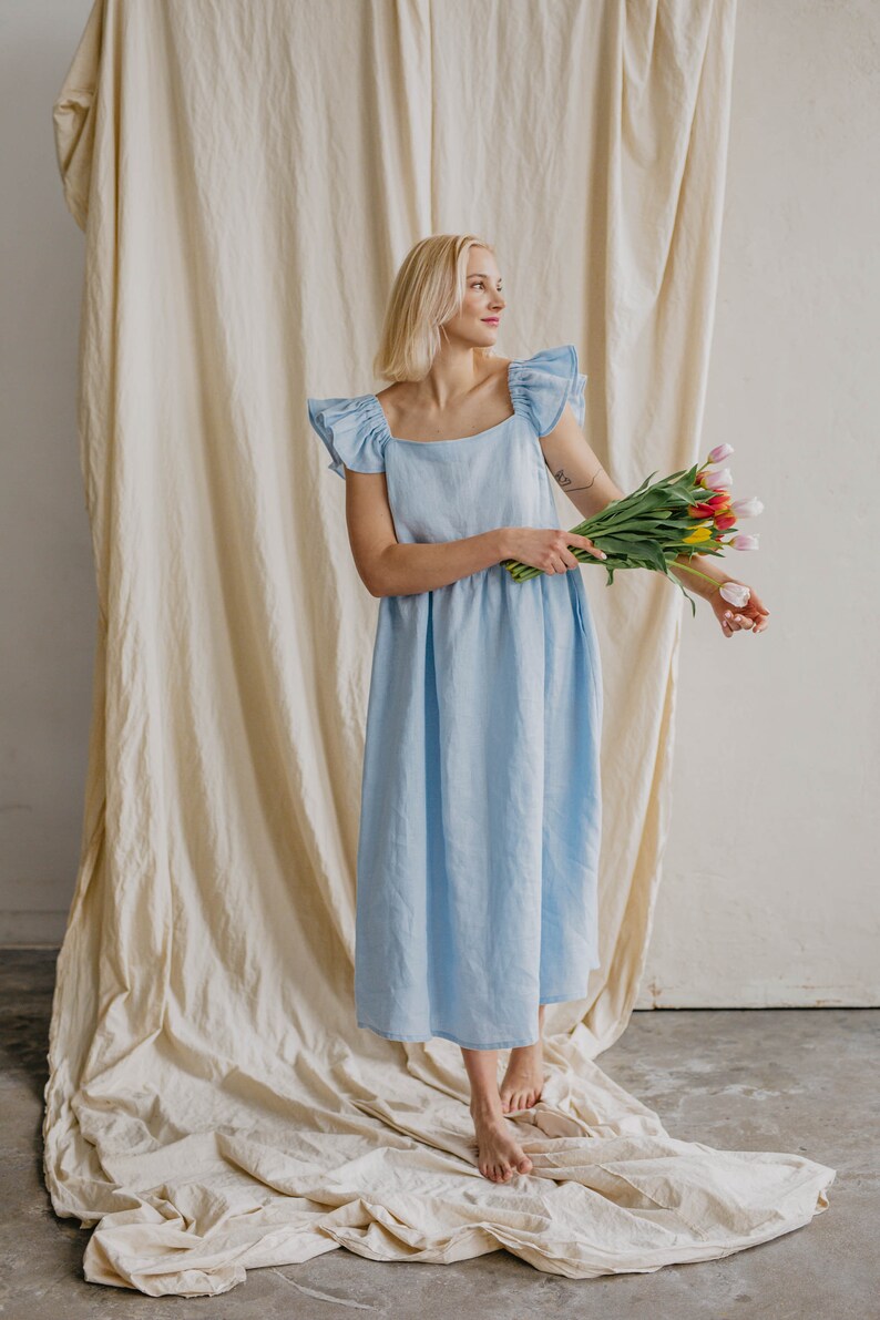 May include: A light blue midi dress featuring a square neckline and ruffled cap sleeves. The dress has a relaxed, loose-fitting silhouette that falls below the knees. The garment is made of a solid-colored fabric and is held with a bouquet of tulips.