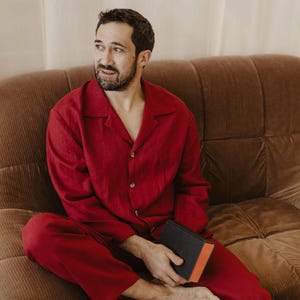 May include: A person wearing a red pyjama set sits on a brown corduroy sofa holding a black and red book.