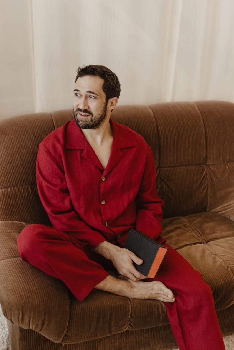 May include: A person wearing a red pyjama set sits on a brown corduroy sofa holding a black and red book.