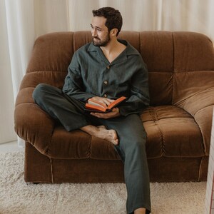 May include: A person wearing a dark green pyjama set sits on a brown corduroy sofa and reads a book. The person is smiling and looking to the right.