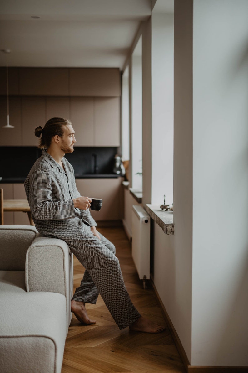 May include: A person in pyjamas sits on a sofa and holds a cup of coffee while looking out a window.
