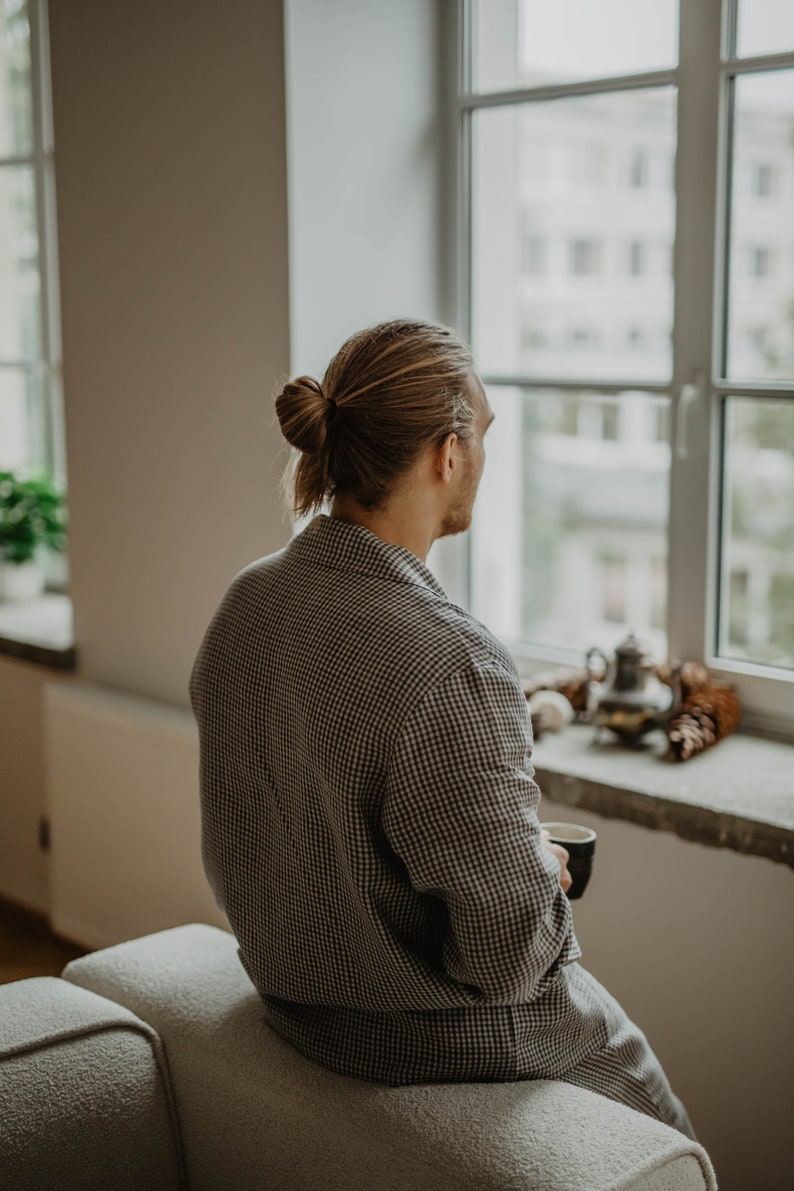 May include: A person wearing a checked shirt sits on a white sofa and looks out a window. The person is holding a cup of coffee or tea.