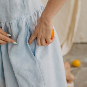 May include: A light blue dress featuring a gathered waistline and a side pocket. The fabric has a visible woven texture, and the garment is shown with a bright orange fruit held near the pocket opening, highlighting the functional design.