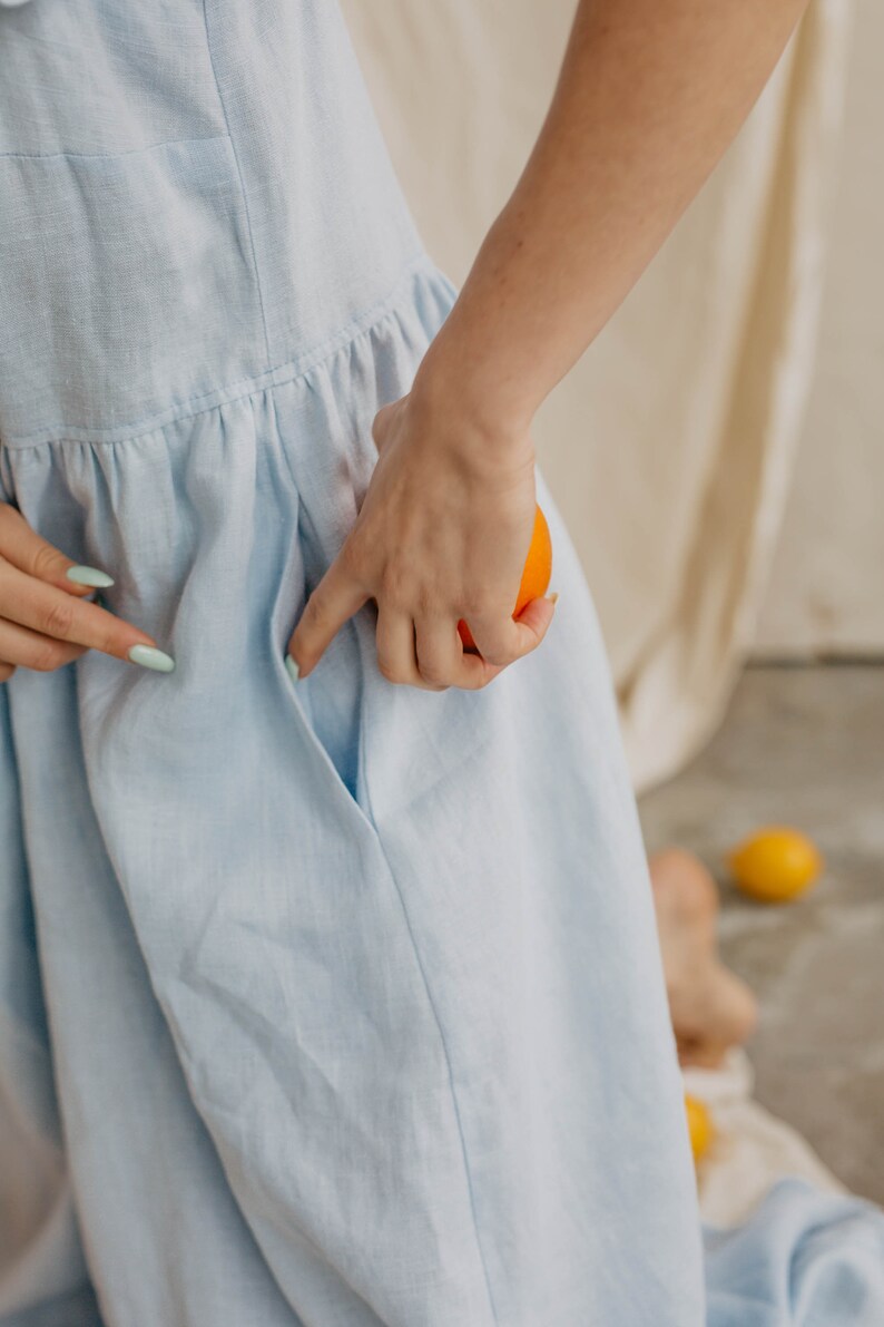 May include: A light blue dress featuring a gathered waistline and a side pocket. The fabric has a visible woven texture, and the garment is shown with a bright orange fruit held near the pocket opening, highlighting the functional design.