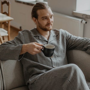 May include: A person wearing a black and white checked pyjama set sits on a sofa and holds a black mug.