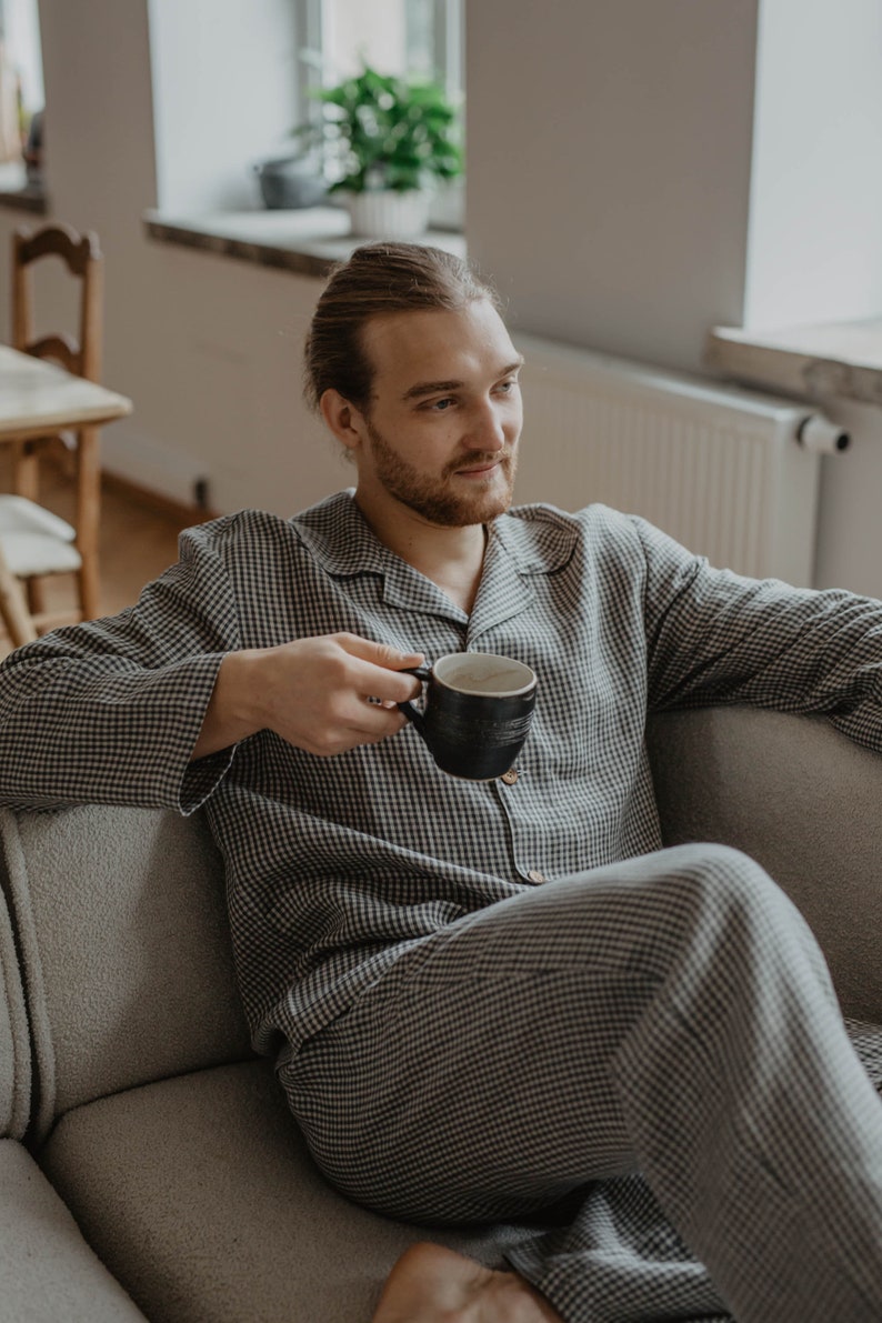 May include: A person wearing a black and white checked pyjama set sits on a sofa and holds a black mug.