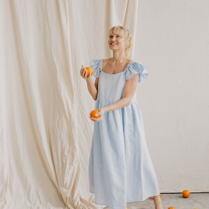 May include: A light blue midi dress with short ruffled sleeves and a square neckline. The garment features a loose, flowing silhouette that reaches mid-calf. Several bright orange citrus fruits are scattered around the floor and in the air.