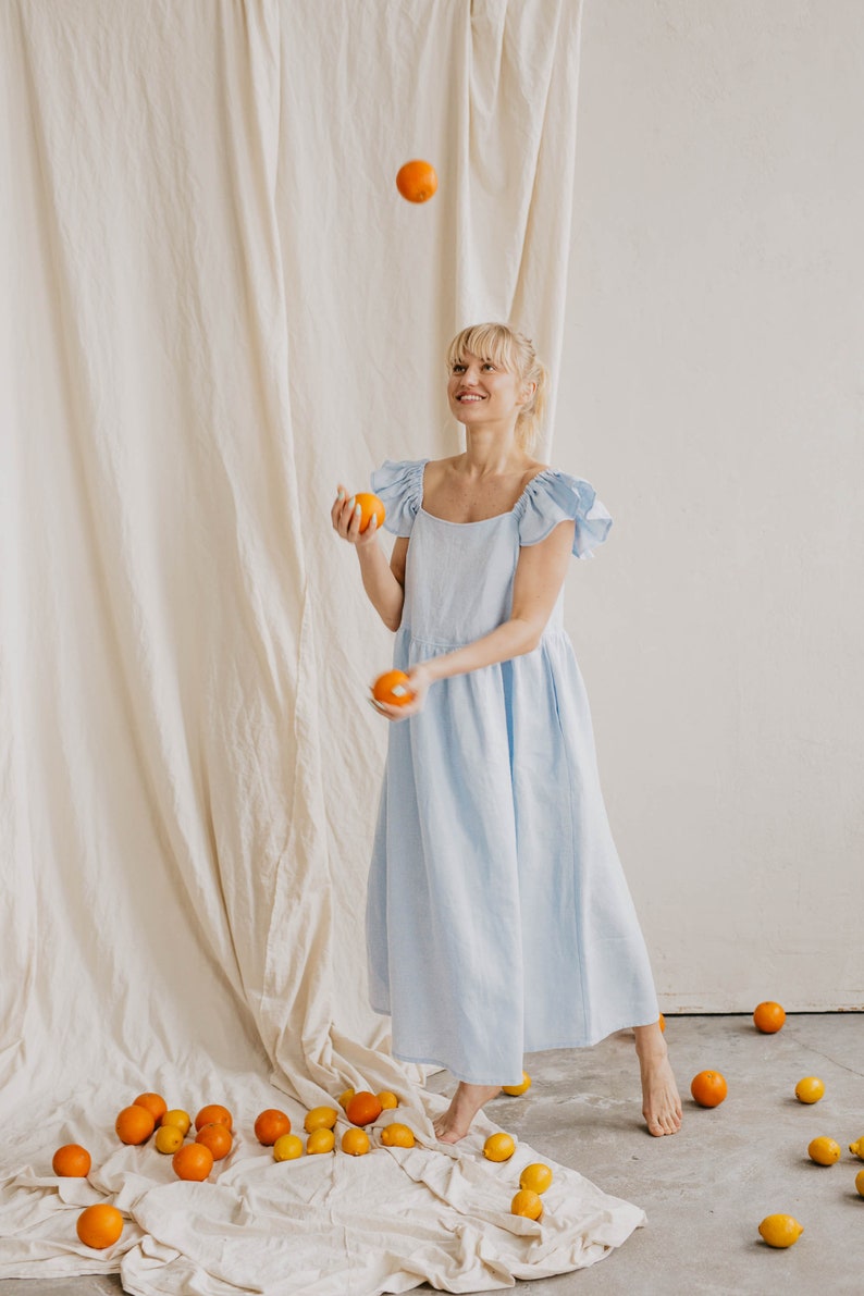 May include: A light blue midi dress with short ruffled sleeves and a square neckline. The garment features a loose, flowing silhouette that reaches mid-calf. Several bright orange citrus fruits are scattered around the floor and in the air.