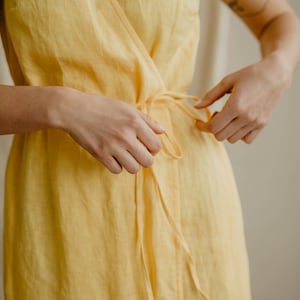 以下が含まれることがあります： ウエストにリボンが付いた黄色のリネン素材のラップドレスです。