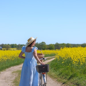 May include: A light blue midi dress with ruffled sleeves and a square neckline, paired with a straw hat featuring a white ribbon. The dress is worn while walking with a bicycle that has a black front basket and a rear wicker basket on a dirt path.