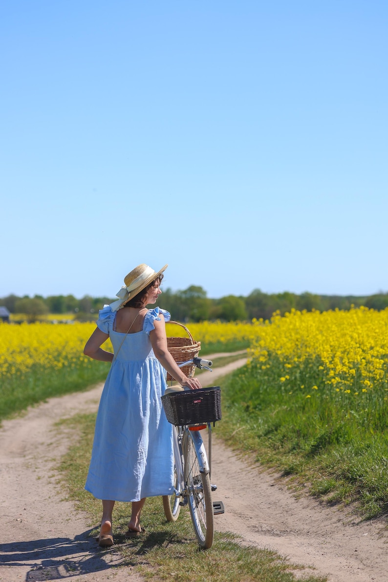 May include: A light blue midi dress with ruffled sleeves and a square neckline, paired with a straw hat featuring a white ribbon. The dress is worn while walking with a bicycle that has a black front basket and a rear wicker basket on a dirt path.
