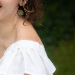 May include: A white off-the-shoulder top with a ruffled neckline and a thin silver chain necklace. A round, textured, muted green earring hangs from the earlobe. The garment features a gathered, elasticized neckline that creates soft, vertical folds.