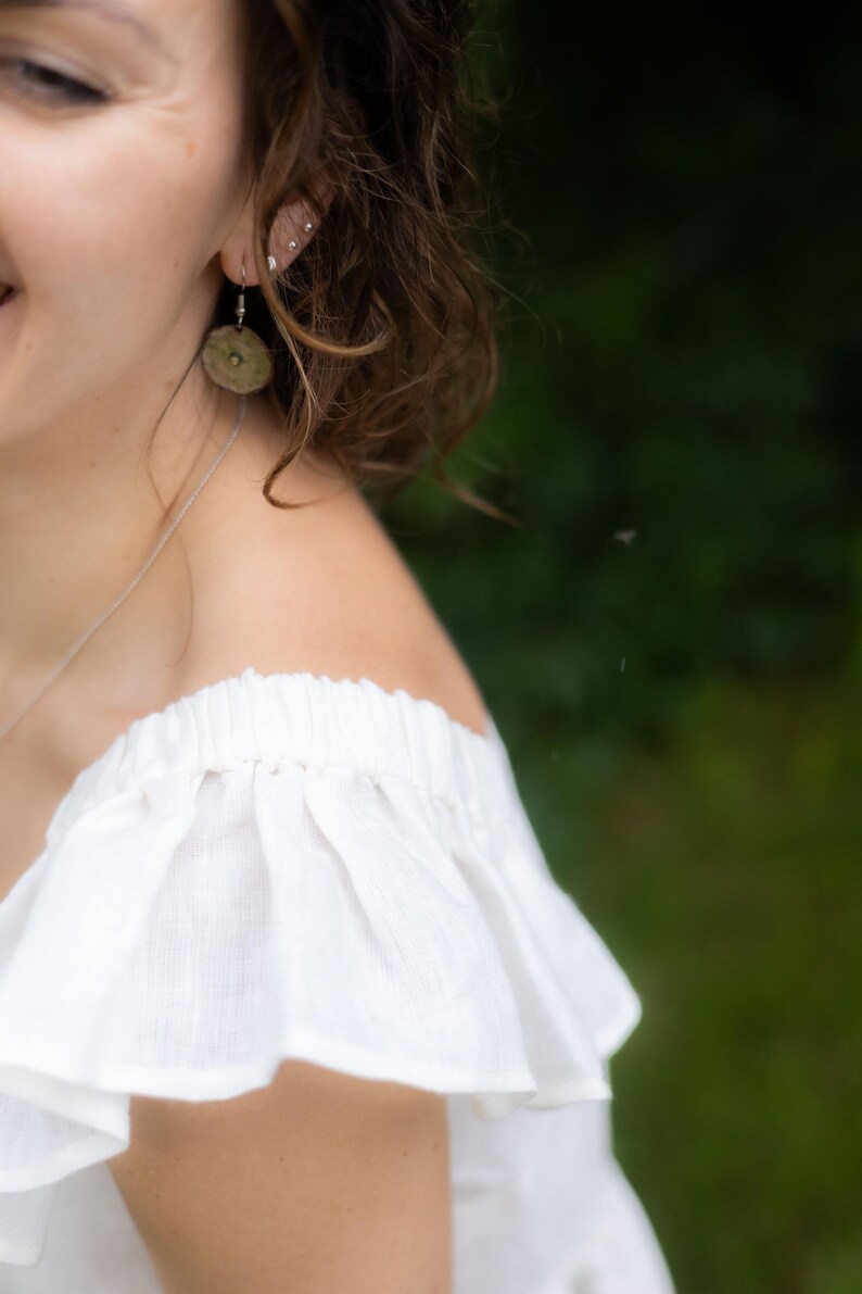May include: A white off-the-shoulder top with a ruffled neckline and a thin silver chain necklace. A round, textured, muted green earring hangs from the earlobe. The garment features a gathered, elasticized neckline that creates soft, vertical folds.