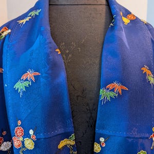 May include: A vibrant blue kimono jacket featuring an intricate woven texture and embroidered motifs of red, white, and yellow flowers alongside green and orange maple leaves scattered across the fabric surface in a repeating pattern.