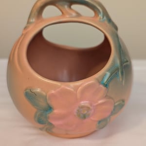 May include: A peach-colored ceramic vase with a large opening and a handle. The vase features a raised floral design in pink and teal. The handle has an open, arched design.