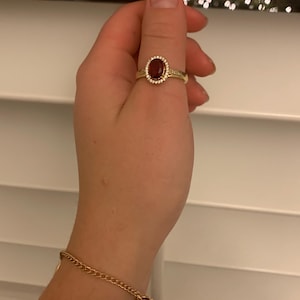 May include: A gold ring with a large oval, dark red gemstone, surrounded by a halo of smaller, clear stones. The ring is on a hand, with a gold chain bracelet visible on the wrist. The background is a white window blind.
