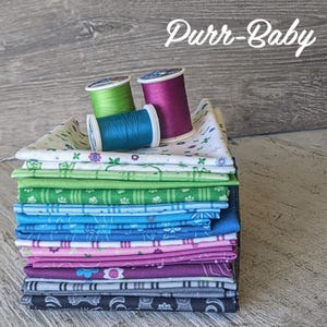 May include: A stack of fabric squares in various colors and patterns, including white, green, blue, pink, purple, and gray. The fabric is arranged in a stack with the white fabric on top. There are three spools of thread on top of the fabric, one green, one blue, and one pink. The text "Purr-Baby" is written in white on a gray background above the fabric.