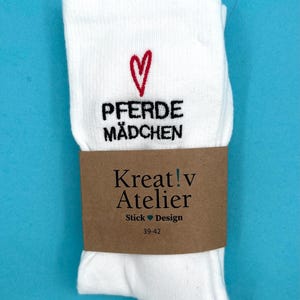 Könnte beinhalten: Weiße Socken mit dem Text "Pferde Mädchen" und einem roten Herz. Die Socken sind mit einem braunen Papierband mit der Aufschrift "Kreat!v Atelier Stick Design 39-42" umwickelt. Auf blauem Hintergrund.