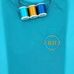 Könnte beinhalten: Ein hellblaues T-Shirt mit einem gestickten Monogramm auf der Vorderseite. Das Monogramm ist ein Kreis mit den Buchstaben "RH" in Gold darin. Drei Garnrollen liegen auf dem Shirt, eine goldene, eine blaue und eine türkisfarbene.