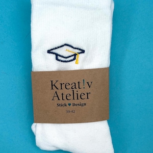 Könnte beinhalten: Weiße Socken mit einer marineblauen und gelben Abschlusskappe-Stickerei. Ein braunes Papierband umschließt die Socken mit dem Text "Kreat!v Atelier" und "Stick Design". Die Socken haben die Größe 39-42.
