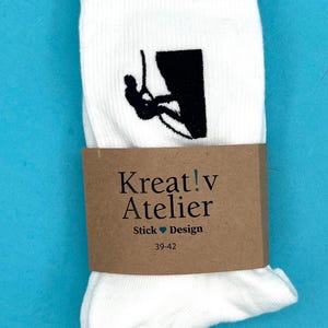 Könnte beinhalten: Weiße Socken mit einer schwarzen Grafik eines Kletterers an einer Klippe. Die Socken sind mit einem braunen Papierband mit der Aufschrift "Kreat!v Atelier Stick Design" und der Größe 39-42 umwickelt.