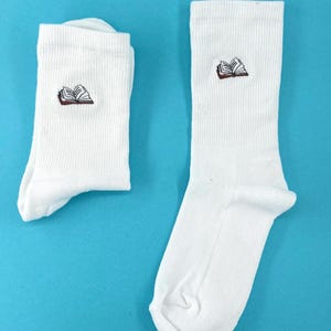 Könnte beinhalten: Ein Paar weiße Socken mit einem gestickten Bild eines offenen Buches auf jeder Socke.
