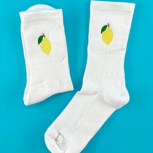 Könnte beinhalten: Zwei weiße Socken mit einer gelben Zitrone auf jeder Socke. Die Zitronen haben grüne Blätter. Die Socken liegen auf einem hellblauen Hintergrund.