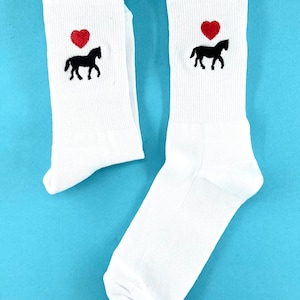 Könnte beinhalten: Ein Paar weiße Socken mit einem schwarzen Pferd und einem roten Herz. Die Socken sind gefaltet und auf einem blauen Hintergrund ausgelegt.