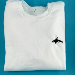 Könnte beinhalten: Weißer Pullover mit einem schwarzen gestickten Orca auf der rechten Brustseite.