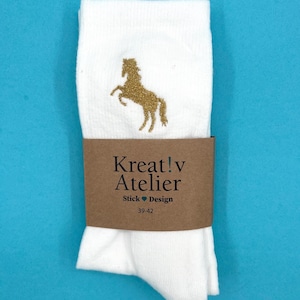Könnte beinhalten: Weiße Socken mit einem goldenen Pferdemotiv. Ein braunes Papierband um die Socken zeigt den Text "Kreat!v Atelier Stick Design 39-42". Die Socken sind aus einem weichen Material gefertigt.