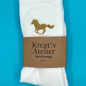 Könnte beinhalten: Weiße Socken mit goldener Pferde-Stickerei. Ein braunes Papierband umwickelt die Socken mit dem Text "Kreat!v Atelier Stick Design" und der Größe "39-42". Die Socken sind vor blauem Hintergrund.