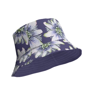 Può includere: Un cappello a secchiello viola con un motivo floreale bianco. I fiori sono di colore lilla chiaro e bianco con accenti verdi.