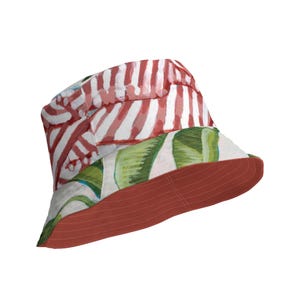 Può includere: Un cappello a secchiello a righe rosse e bianche con una stampa di foglie tropicali verdi e bianche sul bordo. Il cappello è reversibile e ha un lato inferiore rosso pieno.