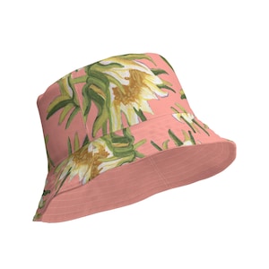 Può includere: Un cappello a secchiello rosa con stampa floreale con fiori bianchi e foglie verdi.