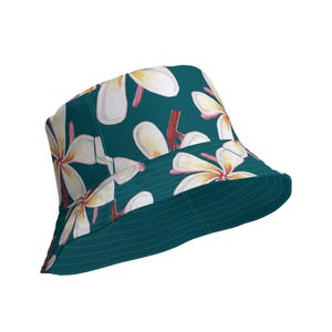 Può includere: Un cappello a secchiello blu-verde con una stampa floreale bianca. I fiori hanno centri rosa e sono disposti in un motivo ripetitivo.