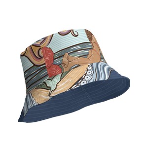 Può includere: Un cappello a secchiello blu con un'illustrazione colorata di una persona sdraiata su una tavola da surf con un polpo e fiori. Il cappello ha un'ampia tesa e una corona piatta.