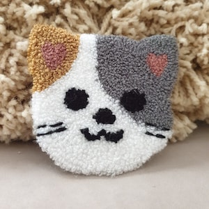 Puede incluir: Una cara de gato blanca, gris y dorada con corazones rosas, bordada en una tela blanca y esponjosa.