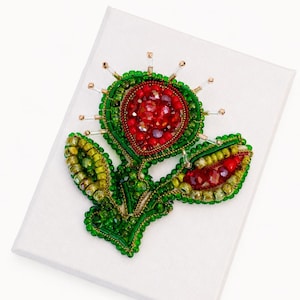 Peut inclure: Une broche perlée faite à la main en forme de fleur stylisée. La fleur centrale présente des perles rouges à facettes entourées de perles de rocaille vert vif et de pointes en verre transparent. Deux tiges feuillues partent de la base.