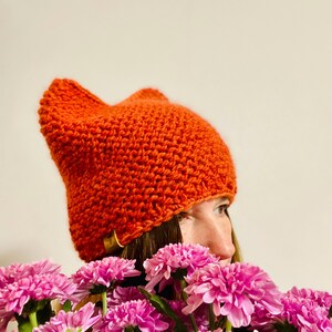 May include: A close-up of a person wearing a bright orange knitted beanie with a unique shape. The beanie is covering the person's forehead and ears. The person is looking to the right, and their face is partially obscured by the beanie. The person is surrounded by a bouquet of pink flowers.
