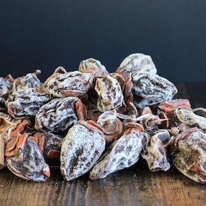 May include: A close-up of a pile of dried persimmons. The persimmons are a dark brown color and have a slightly wrinkled texture. They are arranged in a random pattern on a wooden surface.