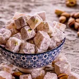 May include: A bowl filled with cube-shaped Turkish delight dusted in white powder, featuring visible green pistachio pieces inside the soft, light purple confection. The bowl has a white base with a blue geometric pattern around the exterior.